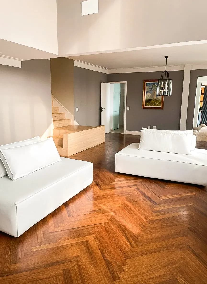 Sala de estar com sofás brancos, piso em madeira espinha de peixe e escada ao fundo